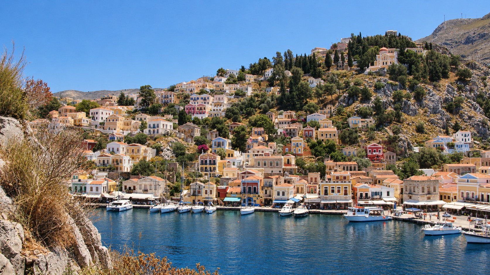 Colourful neoclassical houses climbing a hillside above boats in a Greek island harbour